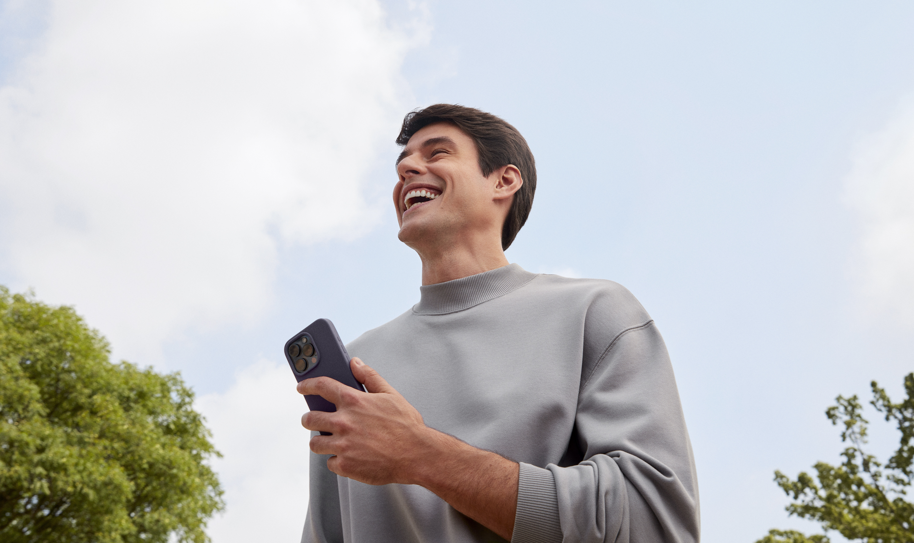 Foto de um homem branco, com uma camiseta cinza de gola alta e um celular na mão. Ele está sorrindo.