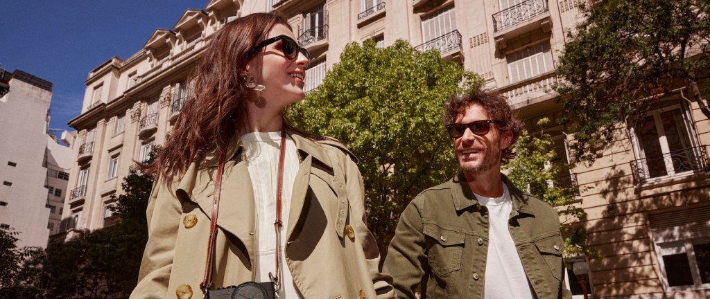 um homem e uma mulher brancos, de cabelos castanhos, caminham entre prédios antigos. Eles usam óculos escuros e vestem casacos verde escuro.