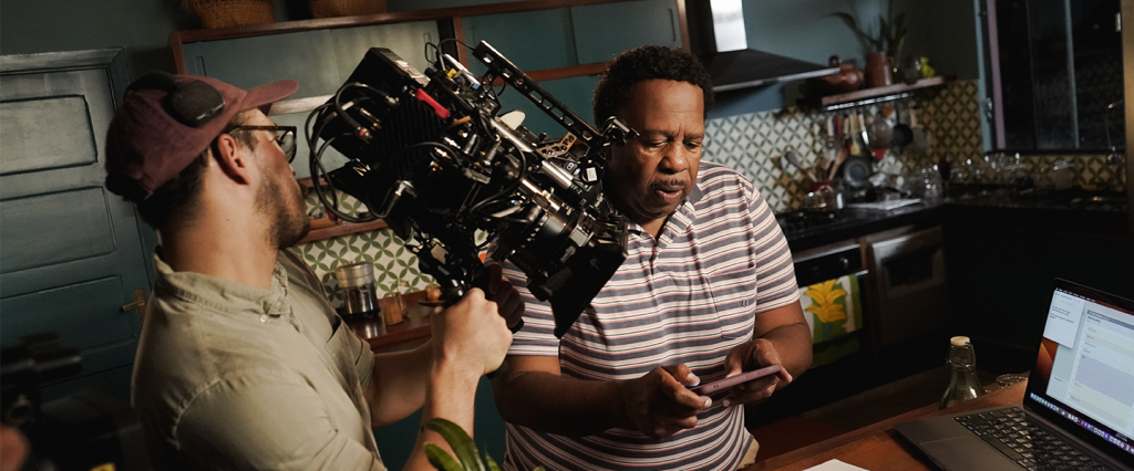 Na imagem, o ator Leslie David Baker aparece concentrado no set de gravação da campanha do Nubank com uma camiseta listrada e olhando para um celular em suas mãos. Na cena aparecem a câmera à esquerda e um laptop à direita.