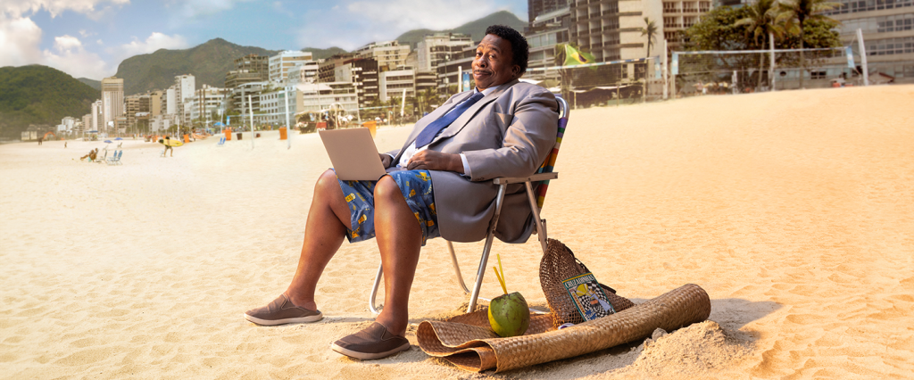 Na imagem, o ator Leslie David Baker aparece na praia de Copacabana sentado em uma cadeira de praia com um laptop no colo vestindo um paletó, camisa e gravata na parte de cima e um shorts de praia na parte de baixo. Ele olha para a câmera com cara debochada.