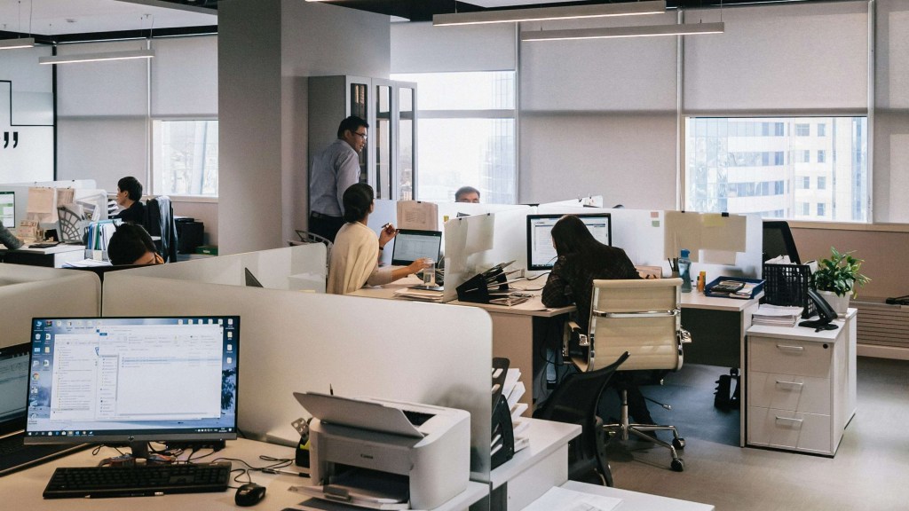 Na imagem aparece a visão geral de um escritório, com um computador ligado à esquerda, uma impressora, algumas baias, papéis, pessoas trabalhando ao fundo, um homem de pé. Parece um cenário de um filme sobre trabalho.