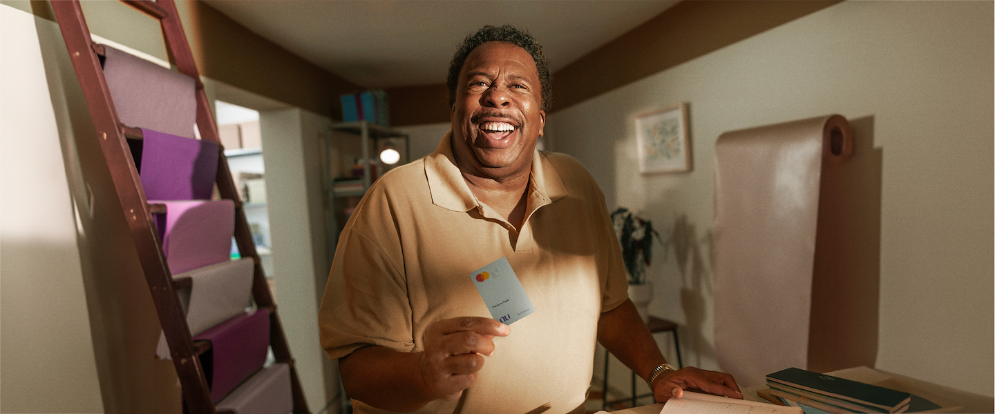 Na imagem, o ator Leslie David Baker aparece sorridente ao centro com uma camisa bege e um cartão PJ do Nubank na mão. Ele está atrás de um balcão de sua papelaria e, à esquerda, se vê blocos de papel em tonalidades de roxo.