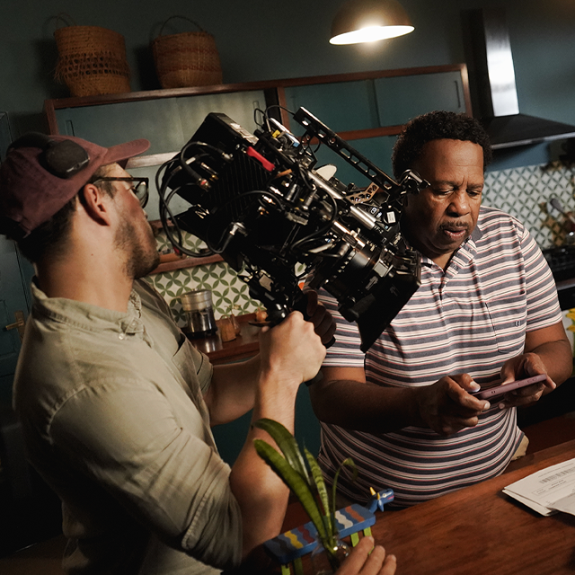Na imagem, o ator Leslie David Baker aparece concentrado no set de gravação da campanha do Nubank com uma camiseta listrada e olhando para um celular em suas mãos. Na cena aparecem a câmera à esquerda e um laptop à direita.