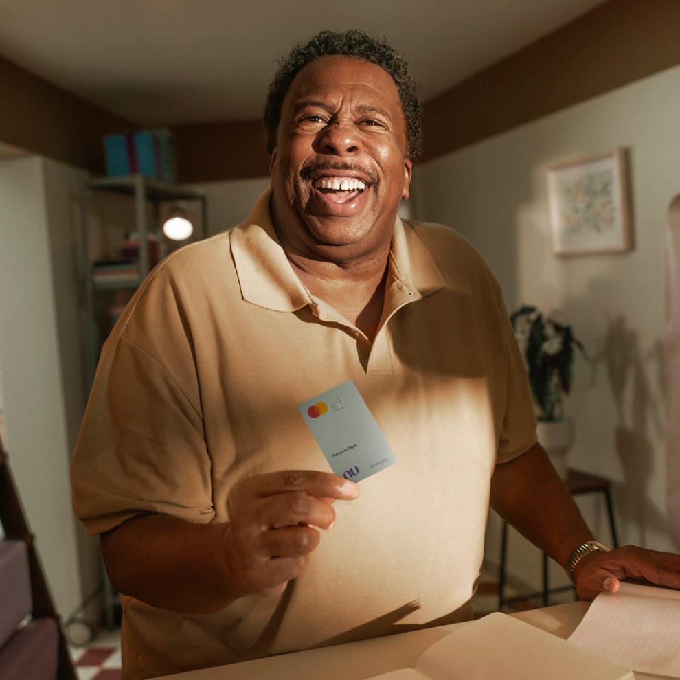 Na imagem, o ator Leslie David Baker aparece sorridente ao centro com uma camisa bege e um cartão PJ do Nubank na mão. Ele está atrás de um balcão de sua papelaria e, à esquerda, se vê blocos de papel em tonalidades de roxo.