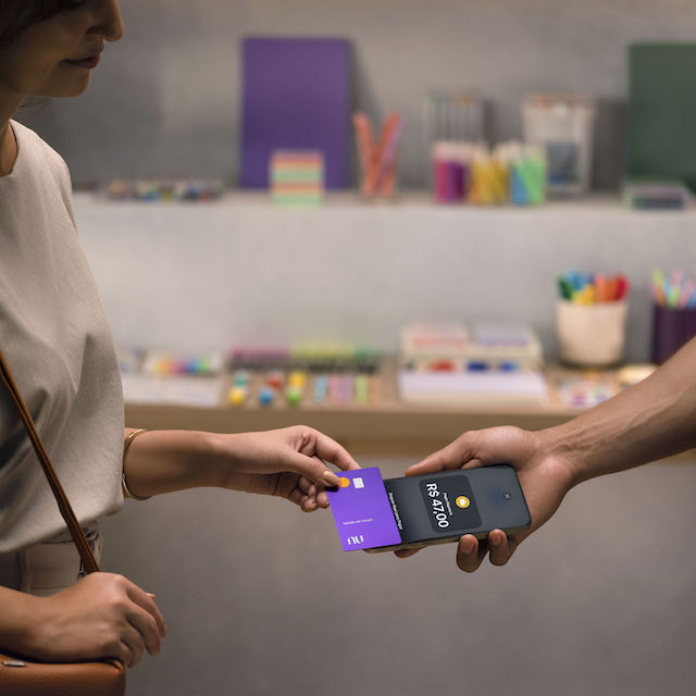 Imagem de uma mão feminina (aparecendo um pouco do corpo também) aproximando um cartão Nubank do iPhone na mão de um homem.