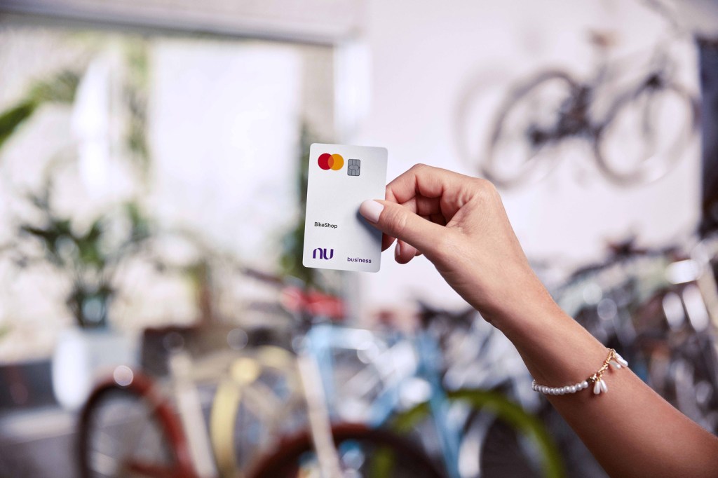 Mão direita de uma pessoa, com pulseira de pérolas no pulso, segura um cartão Nubank business branco. Ao fundo estão várias bicicletas de cores variadas desfocadas.