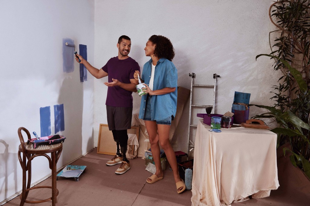 Limite no Nubank - Homem em pé, passa com sua mão direita um rolo de tinta azul em uma parede branca, enquanto olha para a esquerda e conversa com uma mulher negra segurando com a mão esquerda uma lata de tinta branca e com a direita um pincel.