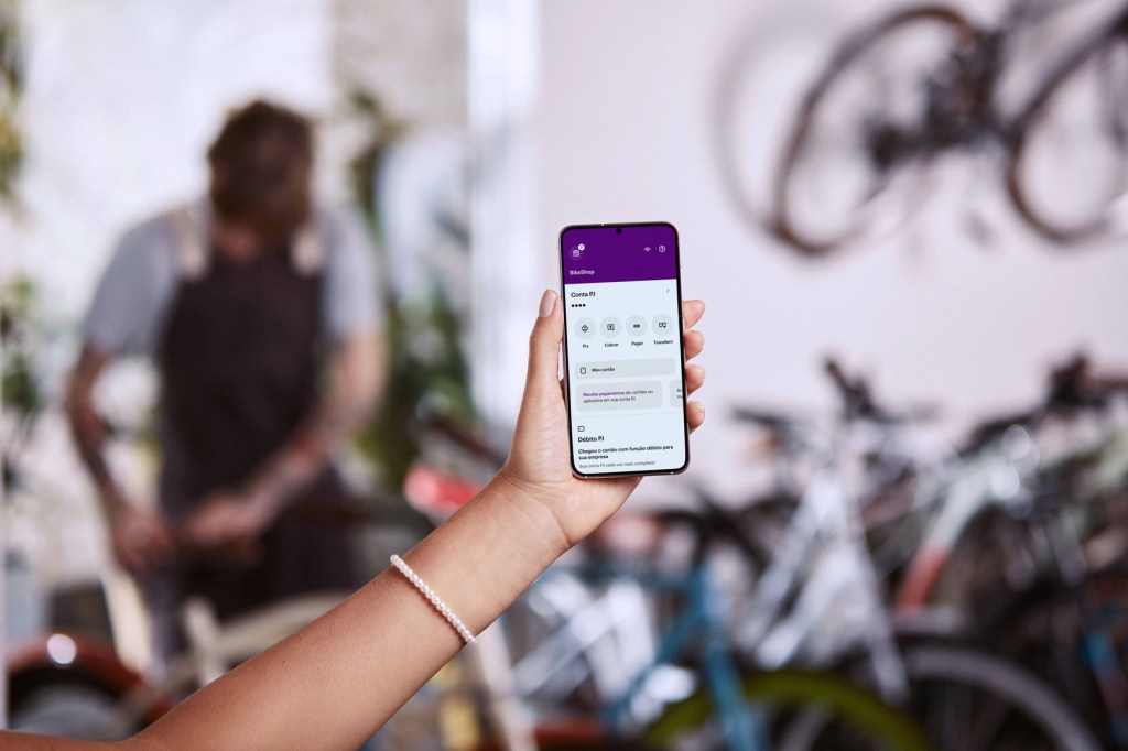 Mão esquerda, com pulseira de pérolas no pulso, segura um celular acessando app Nubank. Ao fundo estão várias bicicletas de cores variadas desfocadas e um homem trabalhando com elas.