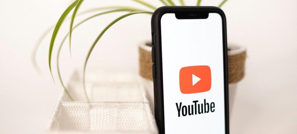 Imagem de um fundo claro. Um celular com a tela do YouTube encostado num vaso de planta e um objeto branco ao lado.