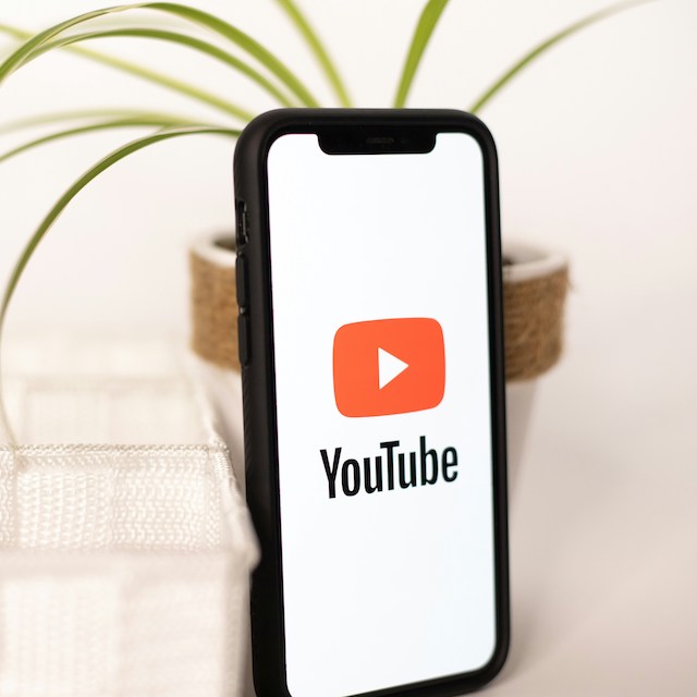 Imagem de um fundo claro. Um celular com a tela do YouTube encostado num vaso de planta e um objeto branco ao lado.