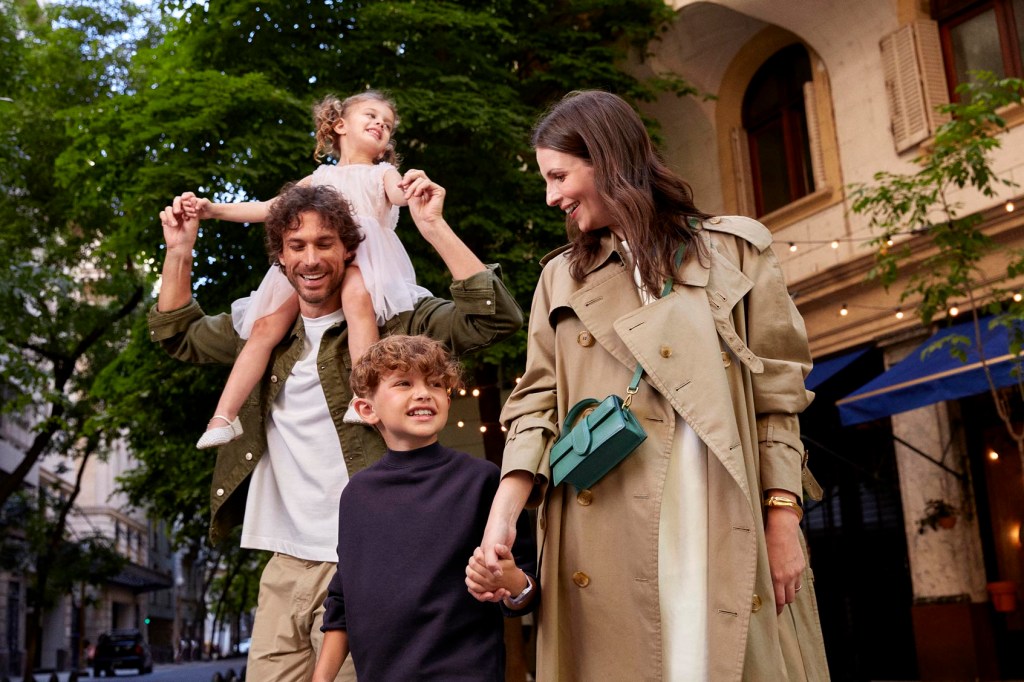 Um grupo que aparenta ser uma família sorrindo e passeando. A mulher está de mãos dadas com o menino e o homem carrega a menina nos ombros. Ao fundo, um sobrado antigo e árvores no meio da cidade.