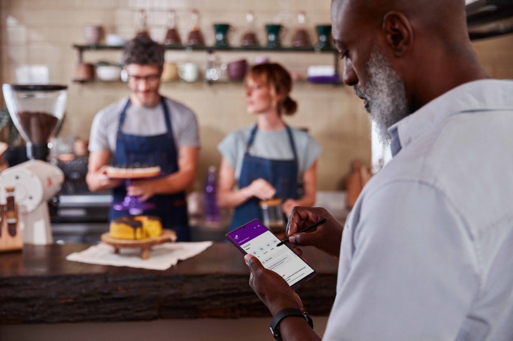 Em primeiro plano, homem negro com barba grisalha mexe na sua conta PJ no App do Nubank. Ao fundo, um homem e uma mulher vestindo uniforme e avental trabalham em cafeteria.