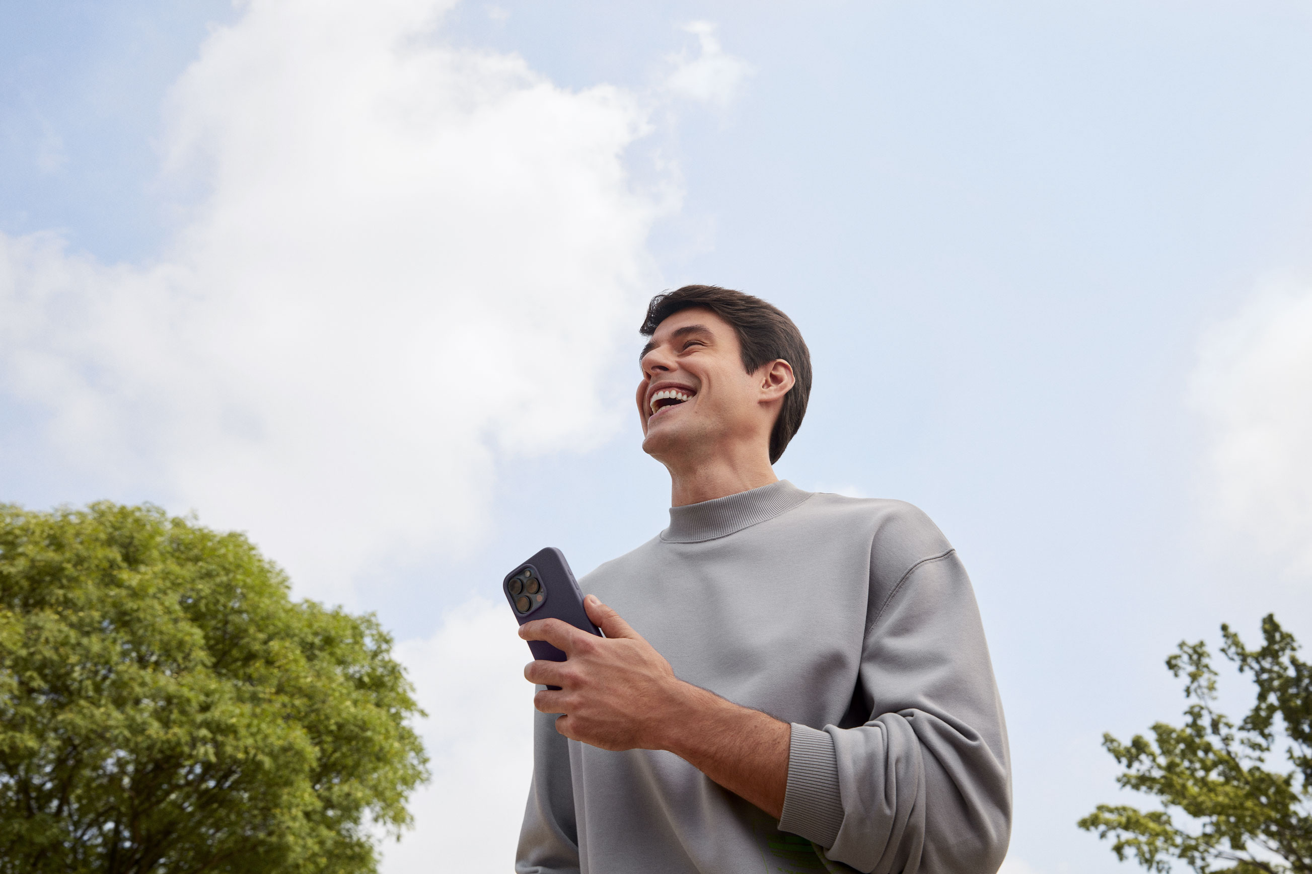 Homem branco sorrindo olhando para frente enquanto segura com a mão esquerda um celular, em um ambiente externo a luz do dia com nuvens no céu e uma árvore a sua esquerda e outra árvore a sua direita.