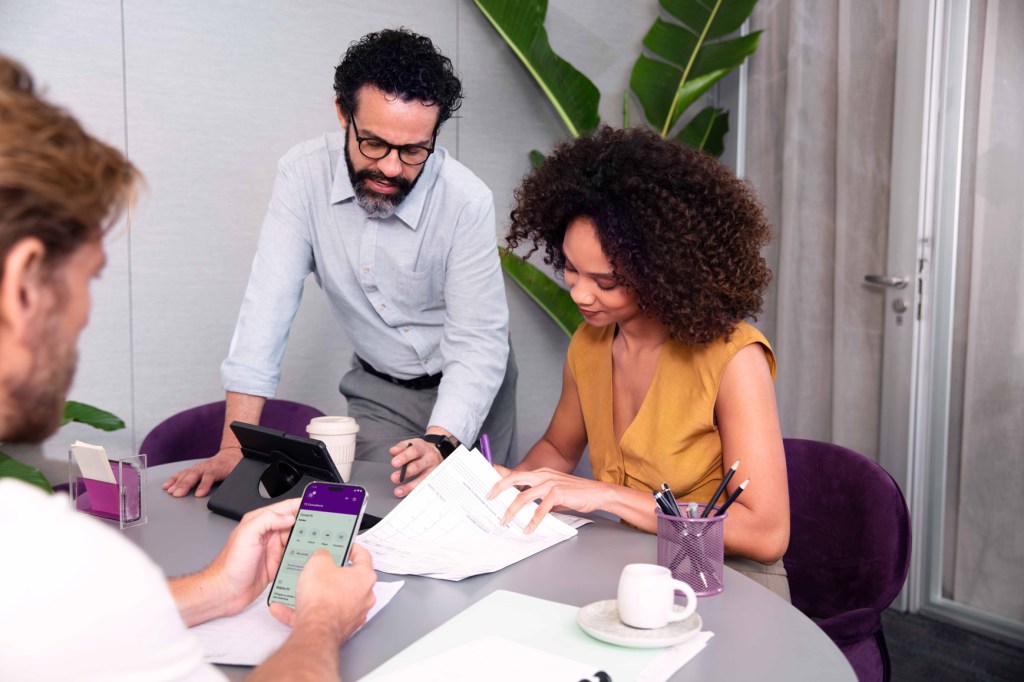 Três pessoas em mesa de escritório. Ao fundo Está um homem em pé, à esquerda, apontando algo em papéis que mulher, sentada, à direita, está lendo. Mais a frente, homem de perfil verifica sua conta no app Nubank.