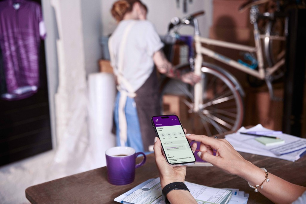 Mãos de uma pessoa aparecem utilizando app Nubank no celular em cima de um balcão com contas, papéis e uma caneca. Ao fundo, de costas e desfocada, mulher trabalha verificando uma bicicleta.
