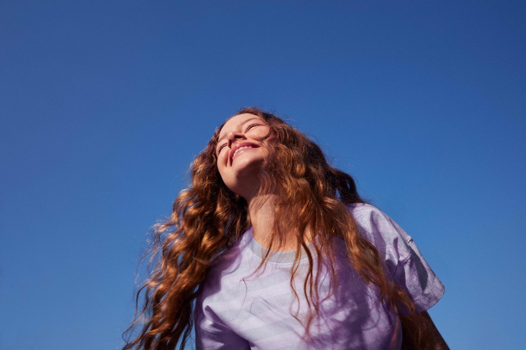 Vista de baixo e do quadril para cima, uma menina branca e ruiva de cabelos cacheados está sorrindo e olhando para frente, em direção ao sol. Céu azul ao fundo.