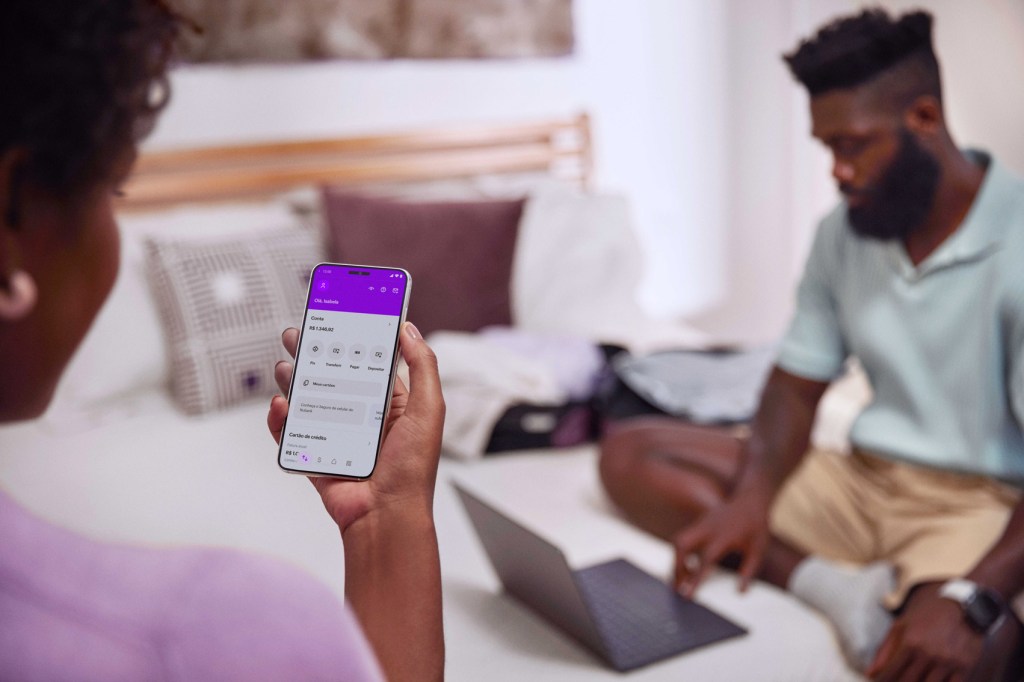 Visão das costas de uma mulher negra segurando com sua mão direita um celular e visualizando sua conta no aplicativo Nubank, ao fundo um homem negro de barba sentado em uma cama de casal e olhando para um notebook.