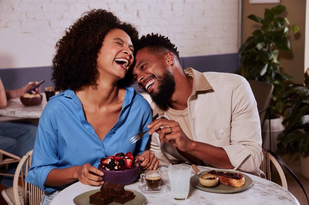 Uma mulher negra à esquerda ri segurando uma tigela com frutas. À sua direita um homem negro ri encostado nela com um garfo em direção à sua tiigela. Ambos estão sentados à uma mesa de café da manhã.