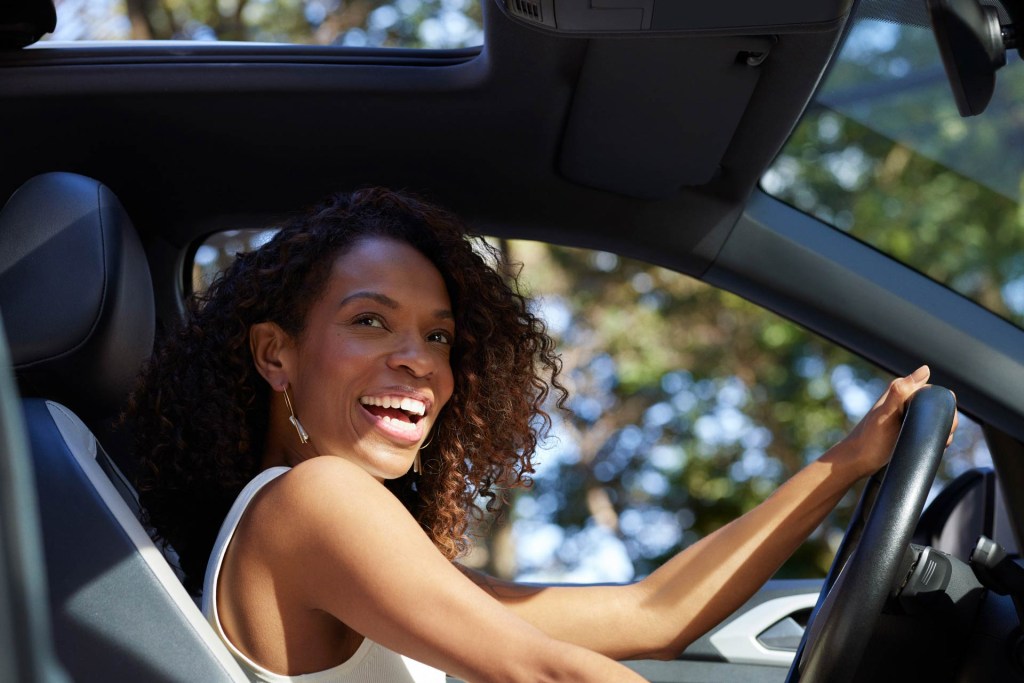  Vista do lado do passageiro de dentro do carro, uma mulher negra de cabelos cacheados está sorrindo com a mão esquerda no volante. Pela janela do carro e desfocada, uma árvore ao fundo.