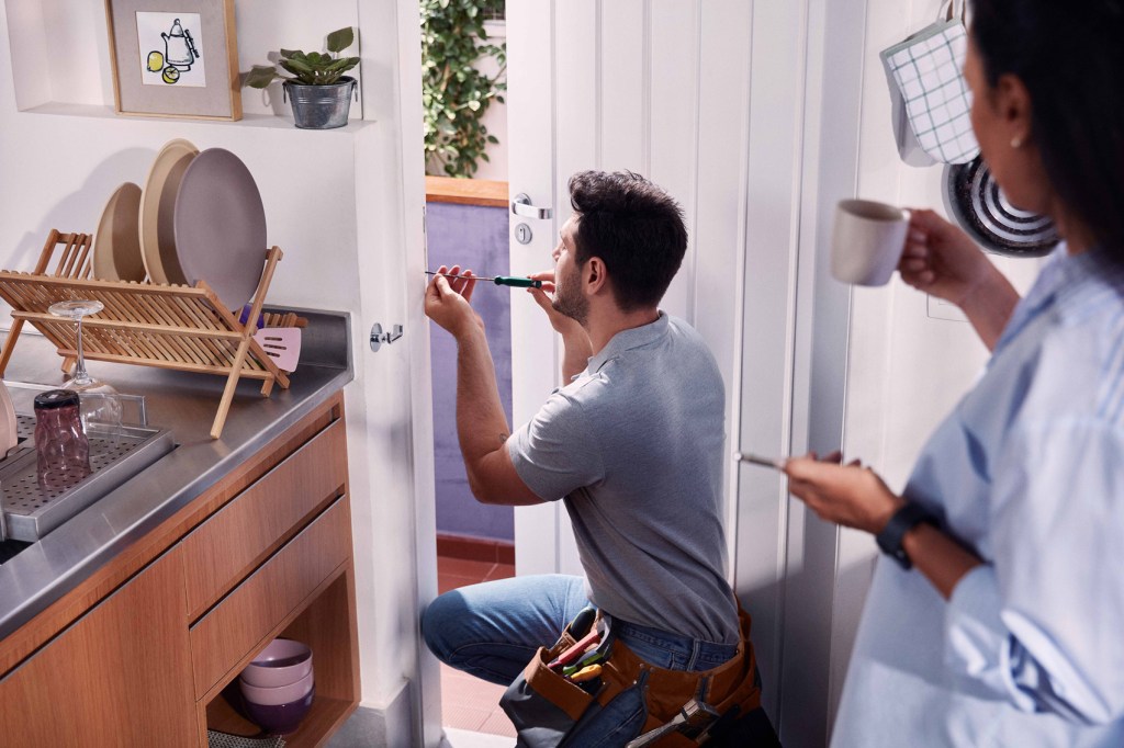 Mulher branca em pé em uma cozinha com um celular na mão esquerda e levando uma xícara à boca com a mão direita, enquanto olha para um homem branco de joelhos consertando uma porta.