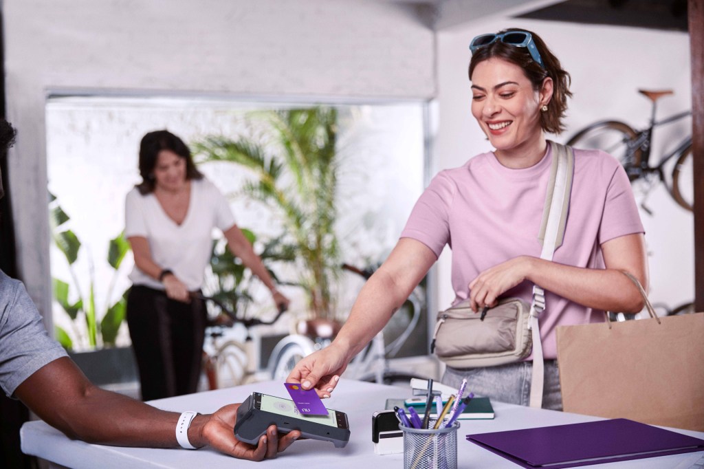 Mulher branca sorrindo usando seu cartão Nubank por aproximação em uma máquina segurada por um braço de um homem negro, ao fundo desfocado uma mulher branca traz uma bicicleta em direção ao visualizador da imagem