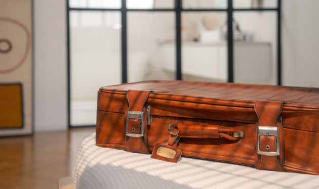 O que é intercâmbio: foto de uma mala de viagem em couro, fechada, em cima de uma cama. O sol invade o ambiente e ilumina a mala.