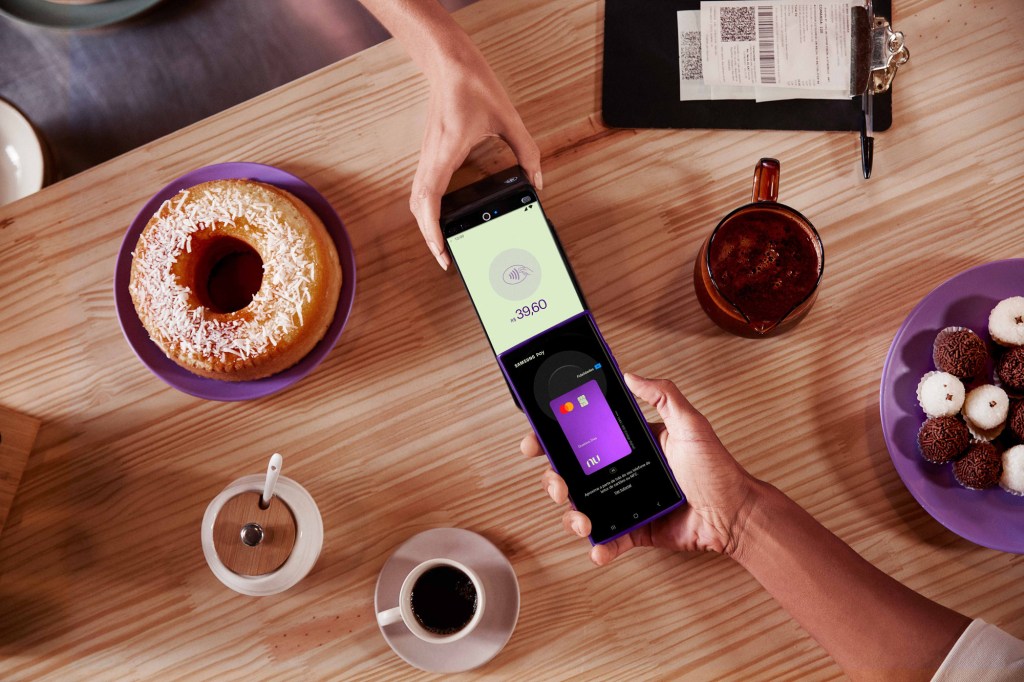 Sobre uma mesa estão um bolo, café, açúcar, brigadeiros e beijinhos, é realizado um pagamento por aproximação utilizando um cartão virtual Nubank no celular.