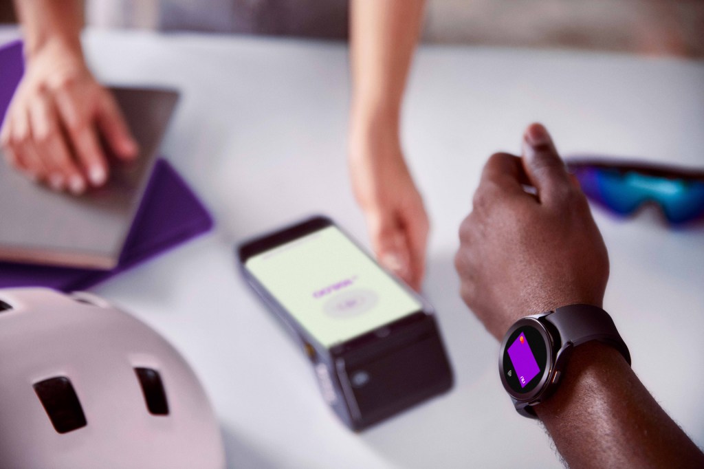 Braço de uma pessoa negra usando um smartwatch para efetuar o pagamento com seu cartão virtual Nubank por aproximação, do outro lado a mão de uma pessoa branca segurando uma máquina de cartão de crédito em cima de uma mesa com um óculos e um capacete de ciclista em cada lado. 