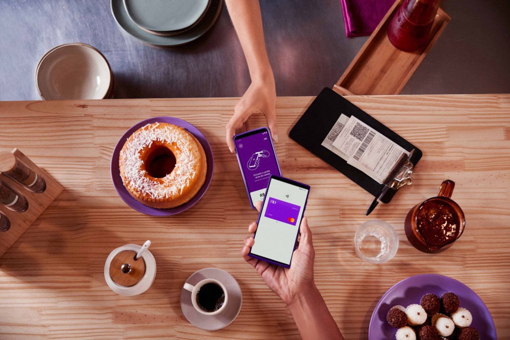 Cartão virtual nubank sendo usado para pagamento por aproximação através do celular. Abaixo está uma mesa com bolo, café e doces.