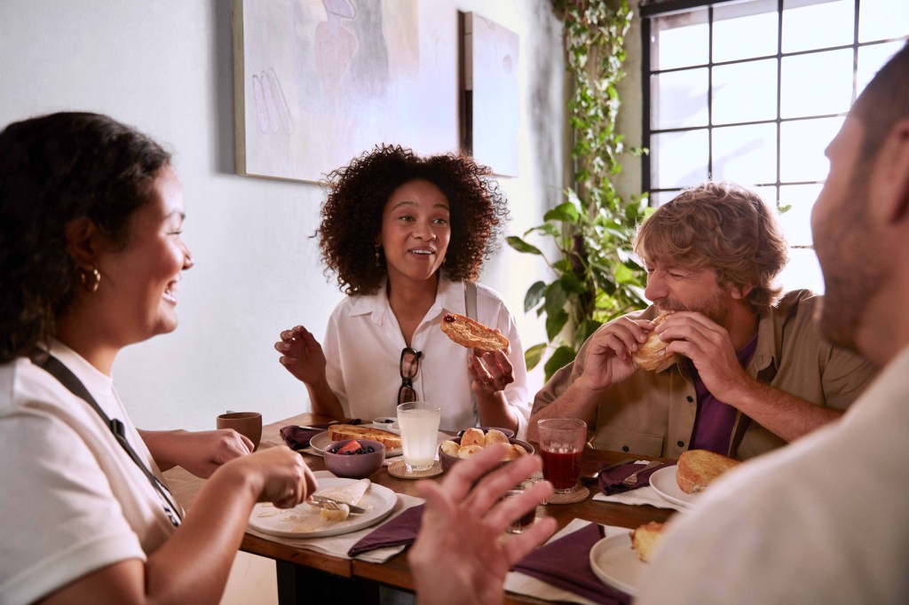 Quatro pessoas, duas mulheres à esquerda e dois homens à direita, sentadas à mesa comendo e rindo. Há dois quadros com pinturas abstratas na parede branca esquerda e, ao fundo, folhagens ao lado de uma janela grande.