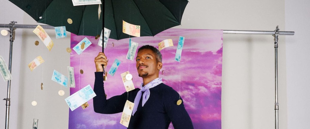 Imagem de um homem negro segurando um guarda-chuva preto, caindo notas de dinheiro do alto. Ao fundo, uma parede com tecido em tons de roxo. Ele usa blusa de frio preta.