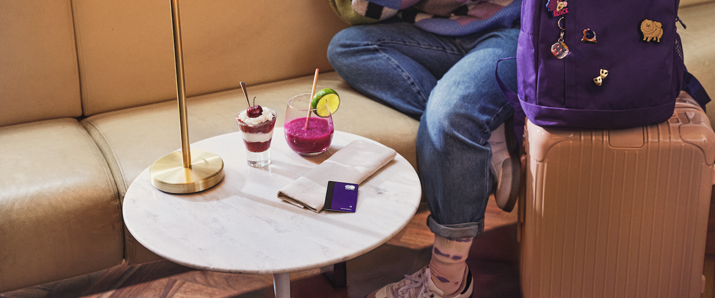 Imagem de uma mesinha de Sala VIP de aeroporto, com docinhos em cima e um cartão Nubank Ultravioleta. Ao lado, uma mala de mão cor rosé com uma mochila roxa em cima. Aparece, sentada no sofá, as pernas de uma menina, usando calça jeans e tênis.