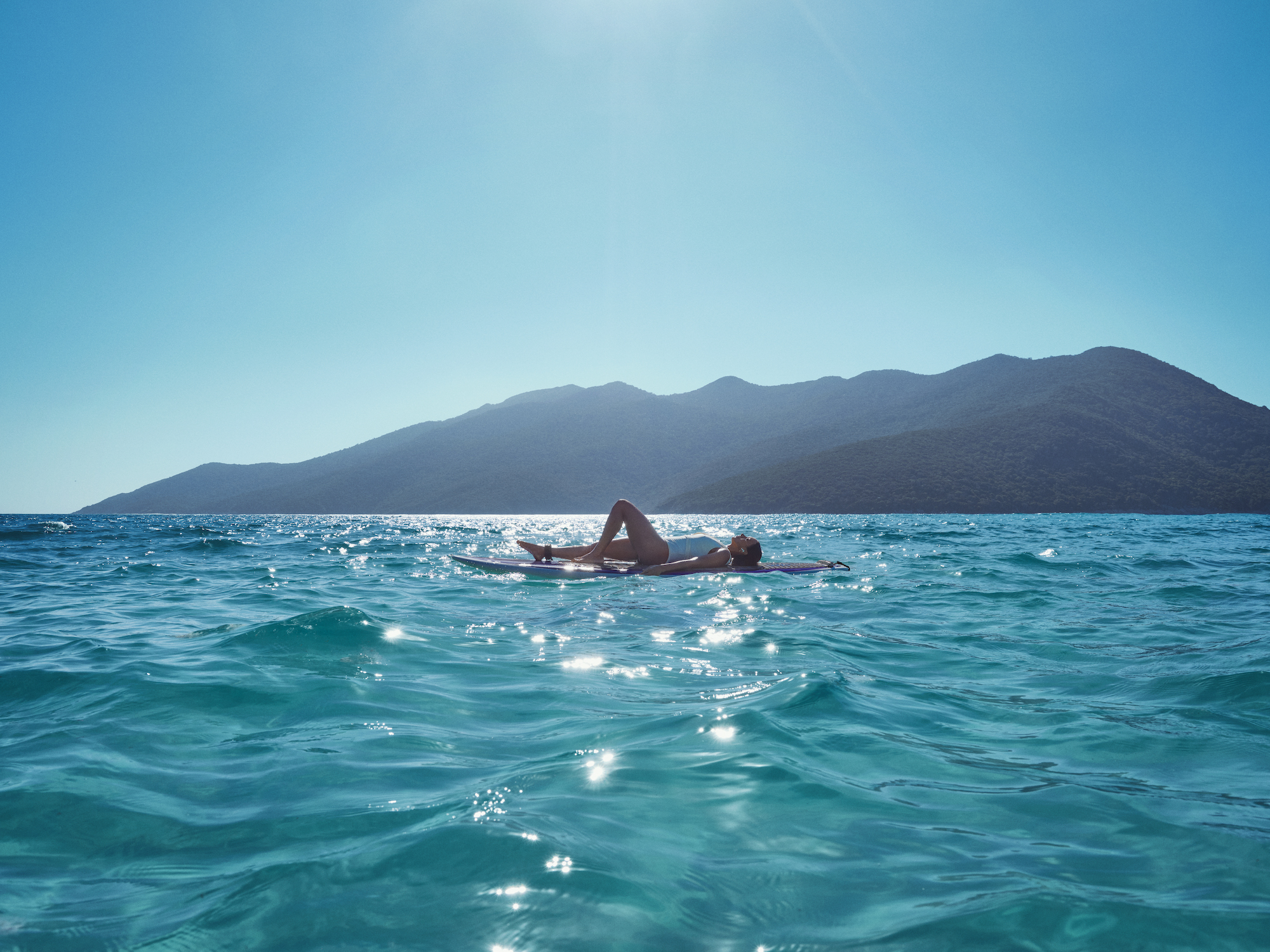 Imagem de uma mulher deitada numa prancha de surf roxa no meio de um mar azul num dia de sol. Ela uso maiô branco.