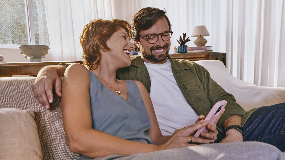 Imagem de uma mulher ruiva de cabelos curtos ao lado de um homem de meia idade, usando óculos. Ela está com o celular na mão e sorrindo. Eles estão sentados no sofá.