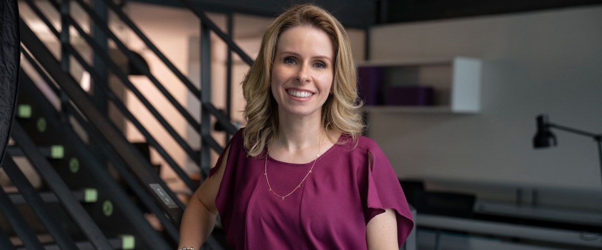 Fotografia de Livia Chanes, CEO do Nubank no Brasil. Livia é uma mulher branca, de cabelos loiros na altura do ombro. Ela tem olhos claros e está sorrindo. Veste uma camiseta cor de vinho e um colar dourado. Ela está encostada na escada do escritório do Nubank, em São Paulo.