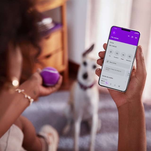 Modo Rua do Nubank para investimentos: imagem de uma mulher segurando um celular enquanto brinca com um cachorrinho. Na tela do celular, é possível ver o menu inicial do aplicativo do Nubank.