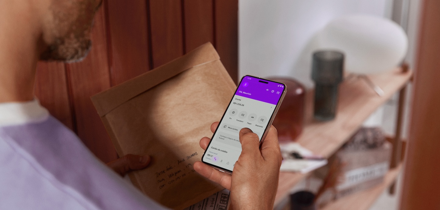 Nubank WhatsApp: imagem de um homem segurando um celular com a tela inicial do app do Nubank aberta. Na outra mão, ele carrega uma correspondência. Ele está parado em frente a uma porta, como se estivesse entrando em casa. A fotografia foi retirada de cima, como se estivéssemos com uma visão sobre os ombros dele.