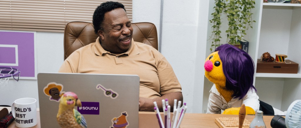 Planos no Papel - Imagem mostra o ator Leslie David Baker sentado em sua mesa no escritório da papelaria Plano no Papel. Ele está ao lado do personagem Adenia, um fantoche amarelo de olhos pretos, nariz rosa e cabelos roxos. Leslie veste uma camisa polo bege. Na mesa é possível ver um computador com adesivos do Nubank, lápis e um papagaio de porcelana.