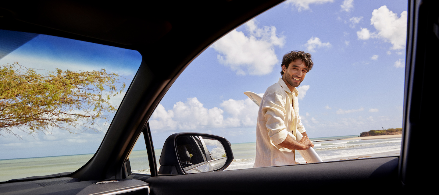 Homem branco, com cabelos dourados cacheados, olha para um carro sorrindo. Ele está na praia e segura uma prancha de surfe.