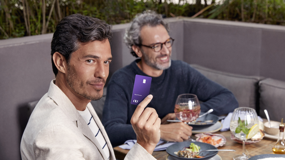 Imagem de dois homens de meia idade, sentados à mesa, com várias comidas e drinks. Um deles está segurando o cartão Ultravioleta.