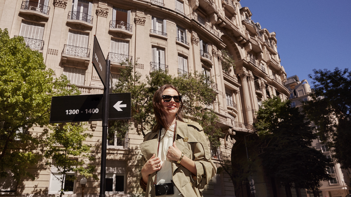 imagem de uma mulher branca de cabelos castanhos sorrindo e usando óculos de sol. Ela está em frente a um edifício antigo.