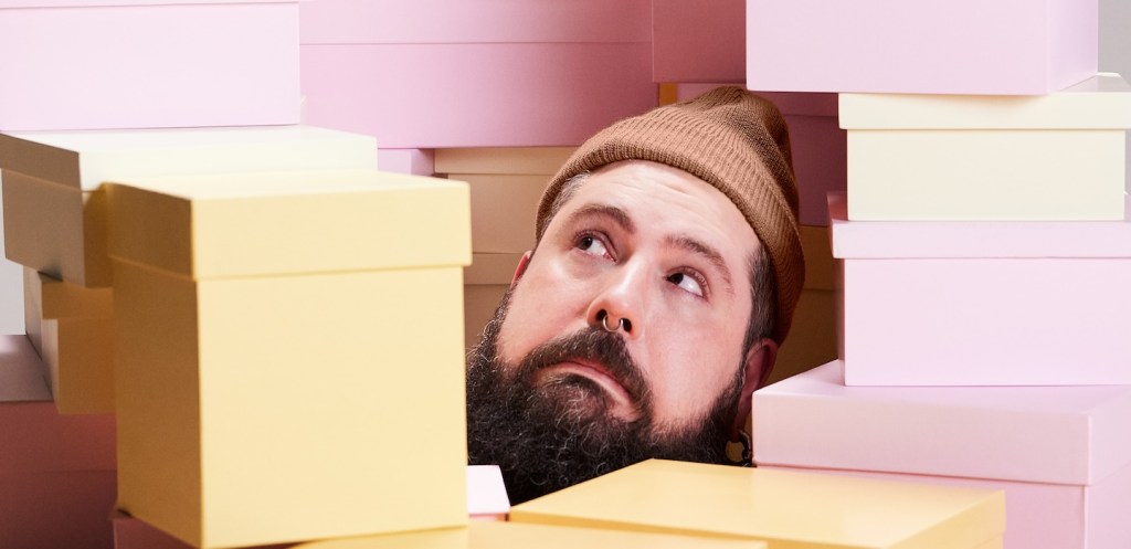 Compras online: imagem de um homem branco preso entre caixas de presente nas cores rosa e amarelo claro. Ele veste uma touca marrom e usa barba.