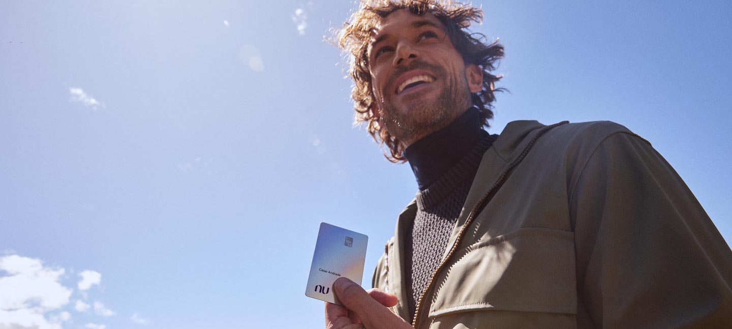 Homem branco, loiro, com cabelo ondulado, está ao ar livre, sorri e segura o cartão multi-moeda do Nubank Ultravioleta, que é branco. Ele veste um casaco verde pesado. O céu está azul.