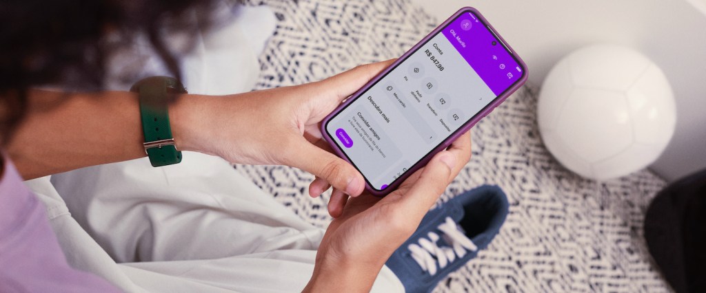 Como comprar Gift Cards pelo app do Nubank sendo menor de 18 anos? Imagem mostra a mão de um adolescente segurando o celular na tela inicial da conta do Nubank para menor de 18 anos. Ao fundo, um tapete cinza e um tênis azul.