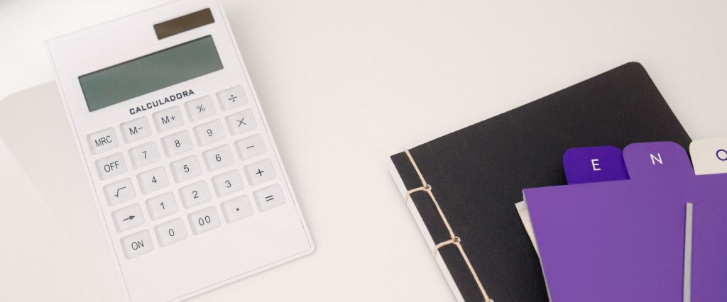 Juros do cartão de crédito - Imagem de uma calculadora branca em cima de uma superfície clara. Ao lado, uma pasta preta e umas fichas em tons de lilás e roxo, com documentos.