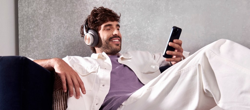 Homem branco, com cabelos ondulados curtos e escuros e barba castanha, está sendo em um sofá olhando para um celular. Ele veste uma camiseta roxa, uma jaqueta e calça brancas e usa fones grandes de ouvido. Ele sorri.