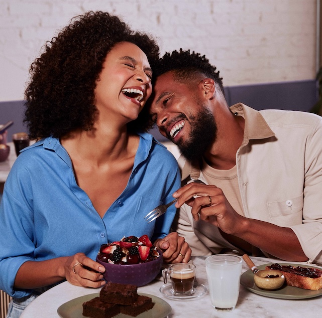 Uma mulher negra, com cabelos escuros cacheados, está ao lado de um homem negro, com cabelos escuros crespos e com barba. Os dois sorriem e estão sentados em frente a uma mesa repleta de frutas.