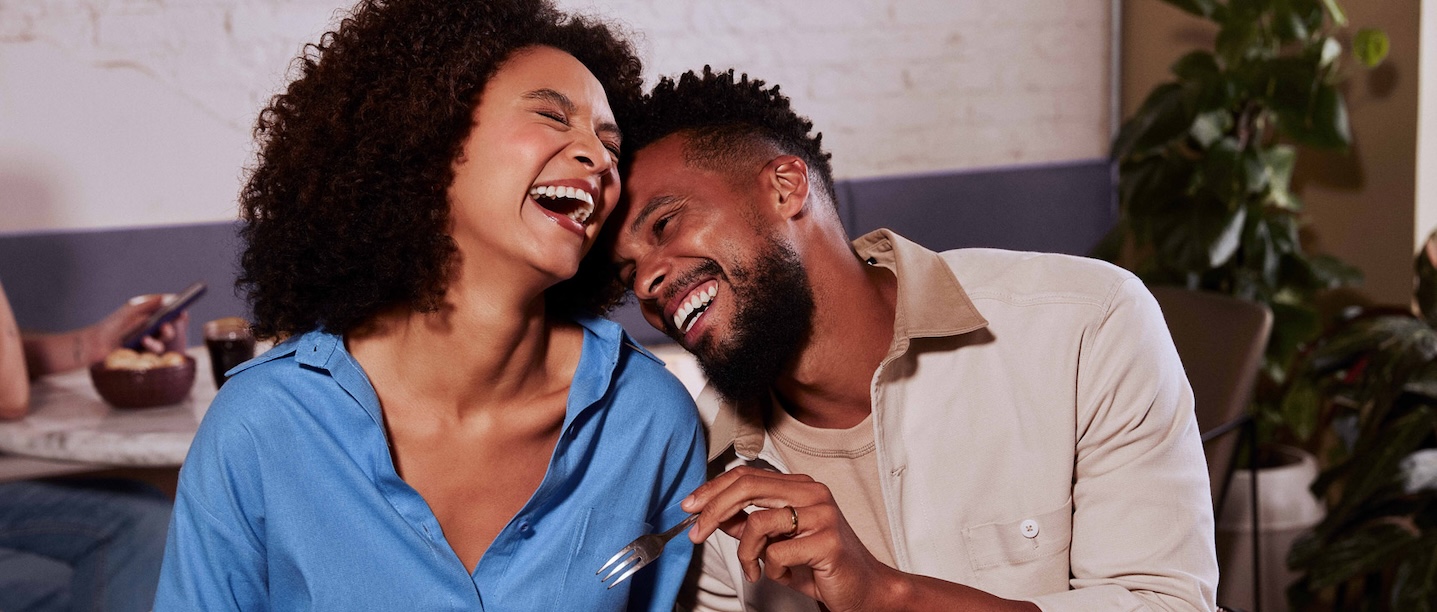 Uma mulher negra, com cabelos escuros cacheados, está ao lado de um homem negro, com cabelos escuros crespos e com barba. Os dois sorriem e estão sentados em frente a uma mesa repleta de frutas.