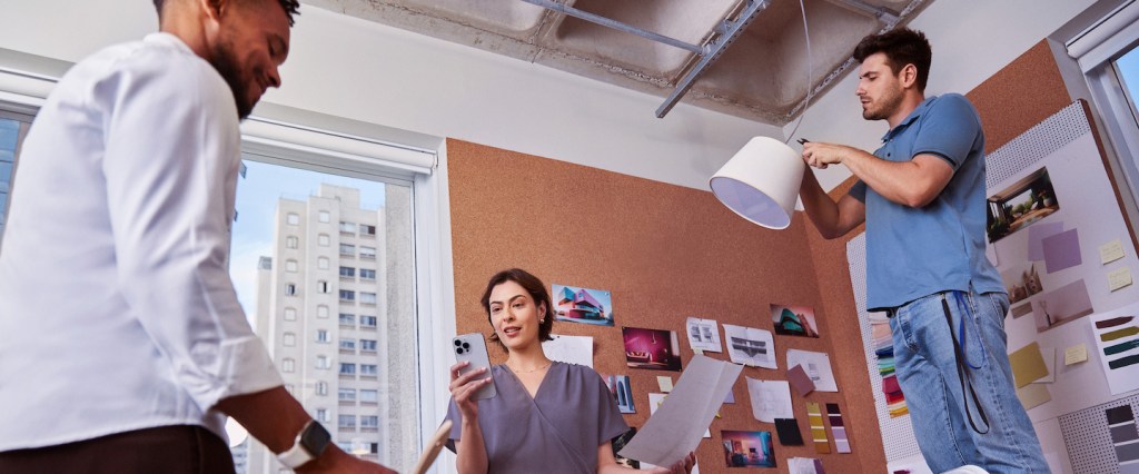 Foto de três pessoas (um mulher no meio e dois homens ao lado dela). Ela está segurando o celular numa mão e uma folha de papel na outra. Eles estão numa sala de trabalho, que tem um painel com informações pregadas e um abajur.