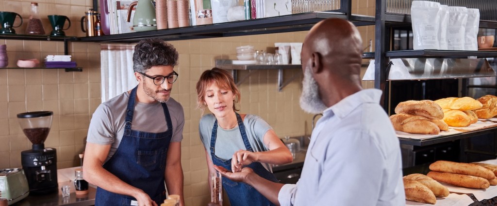 Foto de três pessoas, um homem branco de óculos, uma mulher ruiva e um senhor negro com barba branca. Eles estão em uma padaria, perto de pães e grãos de café.